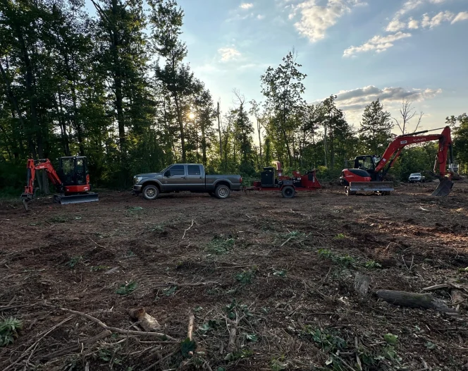 Heavy equipment on cleared land: a grey truck, a wood chipper, and two Kubota excavators, with tall trees.